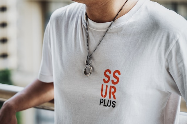 A person is wearing a white t-shirt with the text 'SSUR PLUS' in red and black letters on the front. The individual is also wearing a necklace with a chain and two round pendants. The background appears to have a railing and an out-of-focus cityscape or buildings.