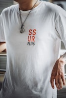 A person is wearing a white t-shirt with red and black text reading 'SSUR PLUS'. A metal chain necklace with a ring hangs from the neck, and the person's right arm is tattooed and resting on a surface.