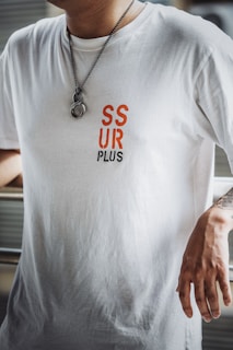 A person is wearing a white t-shirt with red and black text reading 'SSUR PLUS'. A metal chain necklace with a ring hangs from the neck, and the person's right arm is tattooed and resting on a surface.