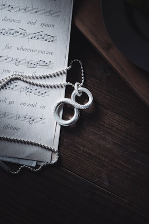 A piece of sheet music lies on a dark wooden surface with a silver chain and two intertwined rings placed on top. The light casts soft shadows, adding depth to the textured wood and highlighting the shiny details of the jewelry.