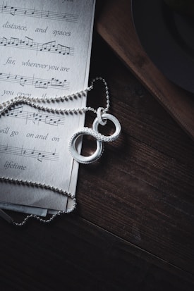 A piece of sheet music lies on a dark wooden surface with a silver chain and two intertwined rings placed on top. The light casts soft shadows, adding depth to the textured wood and highlighting the shiny details of the jewelry.