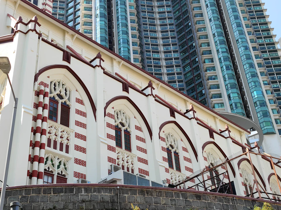 The image shows a large, multi-story building with a white facade and red accents. The building features arched windows on each floor, adding architectural interest to its design. The perspective is from below, looking up at the imposing structure. In front of the building, there are several trees providing greenery to the urban landscape.