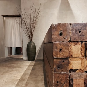 A minimalist interior space featuring a large green vase containing tall, slender branches is placed against a neutral wall. To the right, a stack of heavy wooden beams is visible, showcasing their rugged, natural texture. The floor and walls have an earthy, muted tone, adding to the simplistic and rustic feel.