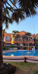 A luxurious outdoor setting featuring a swimming pool surrounded by palm trees and a large, elegant building in the background. The building has an orange facade with arched windows and balconies. Patio furniture is arranged around the poolside, providing a relaxing atmosphere.