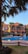 A luxurious outdoor setting featuring a swimming pool surrounded by palm trees and a large, elegant building in the background. The building has an orange facade with arched windows and balconies. Patio furniture is arranged around the poolside, providing a relaxing atmosphere.