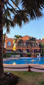 A luxurious outdoor setting featuring a swimming pool surrounded by palm trees and a large, elegant building in the background. The building has an orange facade with arched windows and balconies. Patio furniture is arranged around the poolside, providing a relaxing atmosphere.