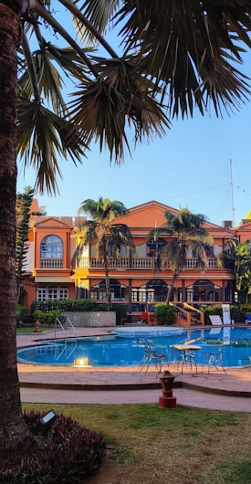 A luxurious outdoor setting featuring a swimming pool surrounded by palm trees and a large, elegant building in the background. The building has an orange facade with arched windows and balconies. Patio furniture is arranged around the poolside, providing a relaxing atmosphere.