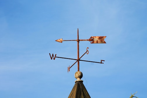 A weather vane atop a pointed roof, featuring cardinal direction indicators with rusted texture, set against a clear blue sky.