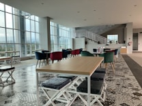 A modern, spacious interior featuring geometric patterned flooring, tables with wooden tops, and colorful chairs, including velvet ones in red and green. Large windows allow natural light to flood the room, and an elegant white staircase leads to an upper level. The room is clean and well-organized, suggesting a cafe or lounge area.