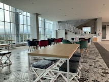 A modern, spacious interior featuring geometric patterned flooring, tables with wooden tops, and colorful chairs, including velvet ones in red and green. Large windows allow natural light to flood the room, and an elegant white staircase leads to an upper level. The room is clean and well-organized, suggesting a cafe or lounge area.