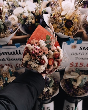 A hand holding a bouquet of dried flowers wrapped in red paper, surrounded by various other dried bouquets displayed for sale with price tags visible.