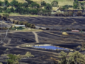 A vast landscape depicting charred fields with visible tractor tracks and a small pond in the foreground. The backdrop has dense green trees contrasting with the scorched land. A farm structure and agricultural machinery, including tractors and a trailer, are situated near the center of the image.