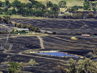 A vast landscape depicting charred fields with visible tractor tracks and a small pond in the foreground. The backdrop has dense green trees contrasting with the scorched land. A farm structure and agricultural machinery, including tractors and a trailer, are situated near the center of the image.
