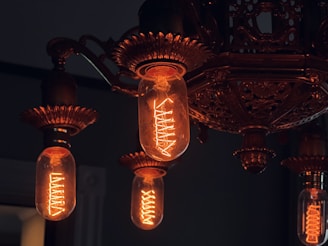 A vintage-style chandelier with multiple glowing Edison bulbs, featuring ornate bronze or brass detailing. The filaments of the bulbs emit a warm, orange glow, creating a cozy and nostalgic atmosphere. The intricate designs of the chandelier add a touch of elegance and antique charm.