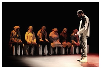 A group of people dressed in muted outfits sits in a dimly lit setting, likely a theatrical performance. One person stands facing them, dressed in similar tones, appearing introspective or solemn.