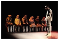A group of people dressed in muted outfits sits in a dimly lit setting, likely a theatrical performance. One person stands facing them, dressed in similar tones, appearing introspective or solemn.