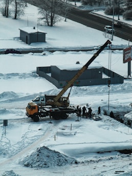A snowy landscape with construction work taking place. A crane truck is positioned on a snow-covered area, lifting or moving something with the help of several workers gathered around. Snow blankets the ground and roofs of nearby small buildings. In the background, there is a road with minimal traffic, and bare trees border the street.
