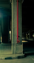 A concrete pillar under a bridge prominently features a red vertical pipe running down its length. The area is dimly lit, creating a stark contrast between the dark shadows and the highlighted parts of the structure. A metal railing surrounds the base of the pillar, and the setting suggests an industrial or urban environment.