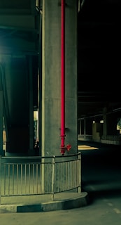 A concrete pillar under a bridge prominently features a red vertical pipe running down its length. The area is dimly lit, creating a stark contrast between the dark shadows and the highlighted parts of the structure. A metal railing surrounds the base of the pillar, and the setting suggests an industrial or urban environment.