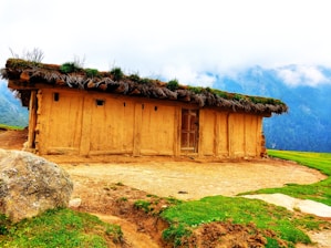 A rustic earthen house with a thatched roof made of grass and plants. The house is set in a scenic mountainous area with mist-covered peaks in the background. There is a large rock and patches of green grass in the foreground, adding to the natural setting.