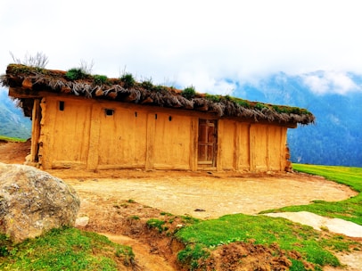 A rustic earthen house with a thatched roof made of grass and plants. The house is set in a scenic mountainous area with mist-covered peaks in the background. There is a large rock and patches of green grass in the foreground, adding to the natural setting.