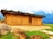 A rustic earthen house with a thatched roof made of grass and plants. The house is set in a scenic mountainous area with mist-covered peaks in the background. There is a large rock and patches of green grass in the foreground, adding to the natural setting.