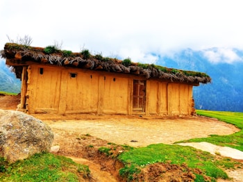 A rustic earthen house with a thatched roof made of grass and plants. The house is set in a scenic mountainous area with mist-covered peaks in the background. There is a large rock and patches of green grass in the foreground, adding to the natural setting.