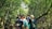 A group of people is walking on a narrow path in a lush, dense forest. The individuals are wearing backpacks and casual clothing, suggesting a nature walk or hike. Sunlight filters through the canopy, casting dappled light on the path and highlighting the vibrant green foliage.