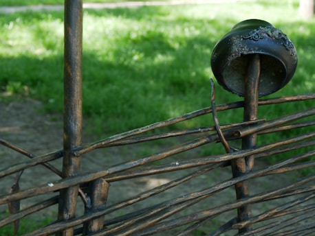 A decorative wrought iron fence featuring a pattern of curved and intertwined metal rods. Sitting atop one of the rods is a weathered, ornate metal cap with detailed embellishments. In the background, there is lush green grass, suggesting a park or garden setting.