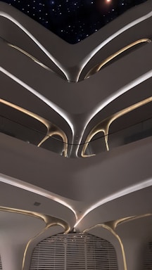 Futuristic interior architecture with multiple floors visible. A series of curving, organic shapes and smooth lines create a futuristic and minimalist aesthetic. Soft lighting highlights these architectural features, with some light sources glowing warmly while others providing cooler illumination. A starry night sky can be seen through an opening at the top, adding to the modern and enigmatic atmosphere.
