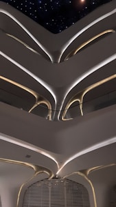Futuristic interior architecture with multiple floors visible. A series of curving, organic shapes and smooth lines create a futuristic and minimalist aesthetic. Soft lighting highlights these architectural features, with some light sources glowing warmly while others providing cooler illumination. A starry night sky can be seen through an opening at the top, adding to the modern and enigmatic atmosphere.