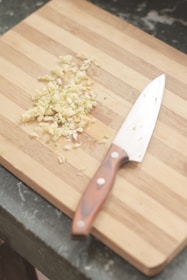 A wooden cutting board with chopped garlic and a chef's knife resting beside it. The grain of the board is visible, and the garlic pieces are scattered in a small pile.