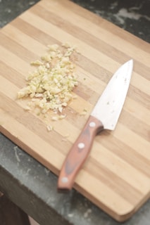 A wooden cutting board with chopped garlic and a chef's knife resting beside it. The grain of the board is visible, and the garlic pieces are scattered in a small pile.