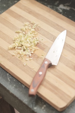 A wooden cutting board with chopped garlic and a chef's knife resting beside it. The grain of the board is visible, and the garlic pieces are scattered in a small pile.