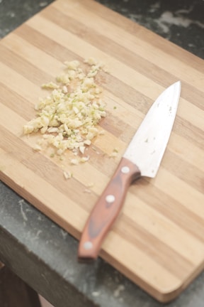 A wooden cutting board with chopped garlic and a chef's knife resting beside it. The grain of the board is visible, and the garlic pieces are scattered in a small pile.