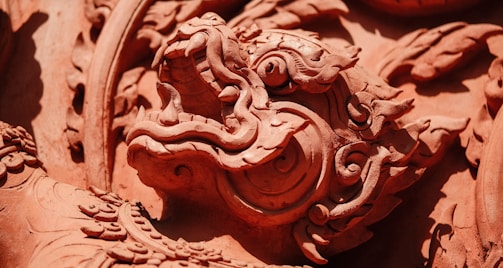 A close-up of an intricately carved dragon or mythical creature in what appears to be terracotta or a similar material. The detailed craftsmanship highlights swirling patterns and sculptural elements, showcasing both the creature's scales and ornate features.