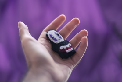 An open hand holds a small, handmade felt figurine with a white face, black body, and red accents, set against a blurred purple background.