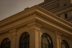 A detailed view of a building corner, showcasing architectural features with arched windows and columns. The structure is composed of light-colored stone and has classical design elements. The building appears to be a multi-story establishment with a flat roof.