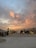A desert setting at sunset, featuring a stunning sky with orange and pink clouds. Tents are pitched on the sandy ground, with several people walking around and a camel being led by a person in the center. The overall atmosphere is lively, with people engaging and moving about.