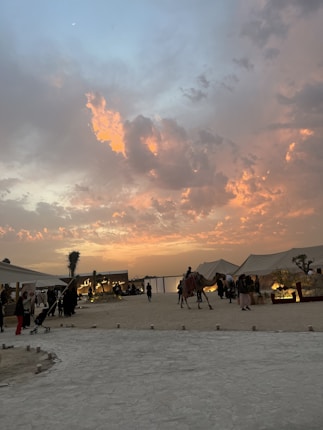 A desert setting at sunset, featuring a stunning sky with orange and pink clouds. Tents are pitched on the sandy ground, with several people walking around and a camel being led by a person in the center. The overall atmosphere is lively, with people engaging and moving about.