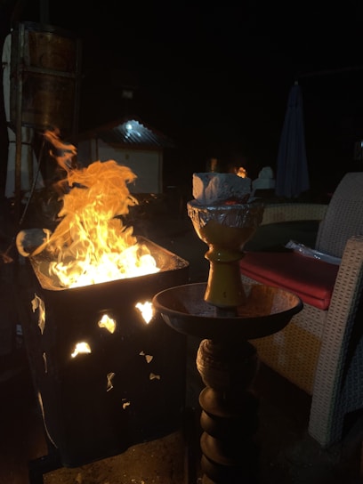 A bright fire is emanating from a metal container with decorative cutouts. In the foreground, there is a hookah with a lit charcoal piece. The scene is set in an outdoor environment with wicker furniture and minimal lighting.