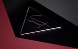 A sleek, triangular black object with reflective surfaces, featuring an engraved metallic signature in the center. The background is primarily a deep red with a shadowed gradient effect, adding depth to the image.