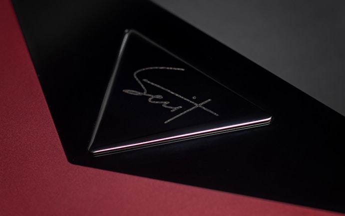 A sleek, triangular black object with reflective surfaces, featuring an engraved metallic signature in the center. The background is primarily a deep red with a shadowed gradient effect, adding depth to the image.