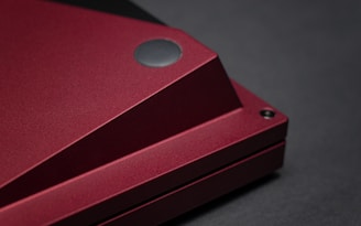 A close-up of a device with a sleek, angular design, featuring a matte dark red finish and a circular black element at one corner.
