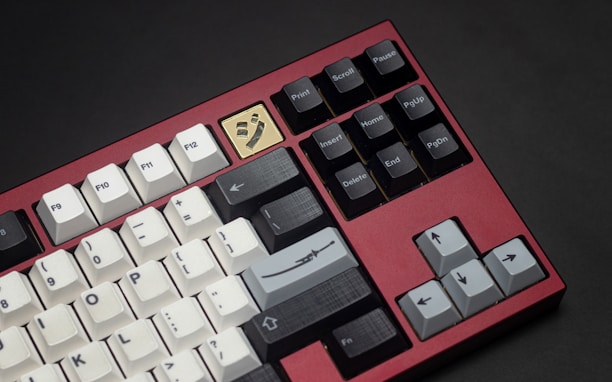 A mechanical keyboard with a red metal frame and a unique golden keycap with an artistic symbol. The keyboard features a mix of white, black, and gray keys, including function keys and arrow keys. The design is sleek and modern, emphasizing both aesthetics and functionality.