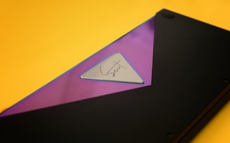 A close-up of a sleek, modern object featuring a triangular metallic element with a reflective surface, which is embedded in a smooth black casing. The background is a vibrant yellow, contrasting sharply with the dark object. The central triangular piece has a signature, adding a personal touch or branding element.