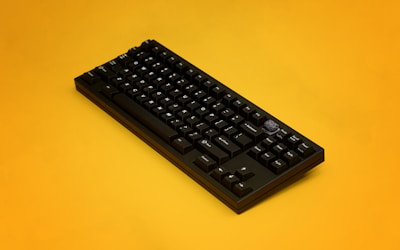 A sleek black mechanical keyboard is positioned on a vibrant yellow background. The keyboard features clean lines and has a single unique keycap with a decorative design.