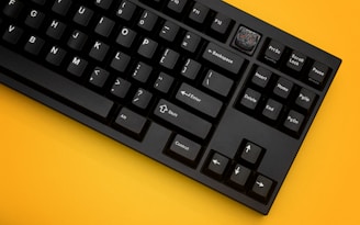 A black mechanical keyboard with white lettering on the keys is placed on a vivid yellow background. One of the keys, located in the top right section, is replaced with a custom keycap featuring a detailed design.