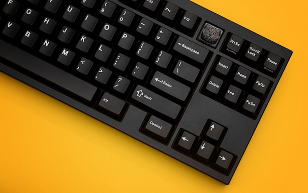 A black mechanical keyboard with white lettering on the keys is placed on a vivid yellow background. One of the keys, located in the top right section, is replaced with a custom keycap featuring a detailed design.