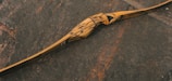 A beautifully crafted wooden bow with intricate patterns lies on a textured stone surface. The bow features a natural wood grain with darker, swirling designs, emphasizing its artisanal quality.